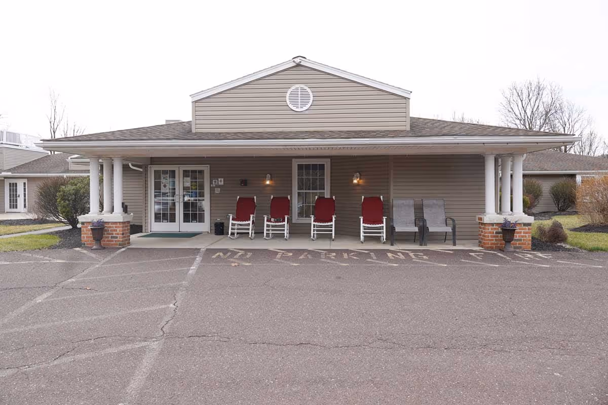 Front exterior view of a single-story senior living facility building with beige siding and a covered porch. The porch has four red cushioned rocking chairs and two gray chairs. There are two white columns on brick bases supporting the porch roof. The ground in front has a 'NO PARKING' sign painted on the asphalt. Some bushes and grass are visible on either side of the building.