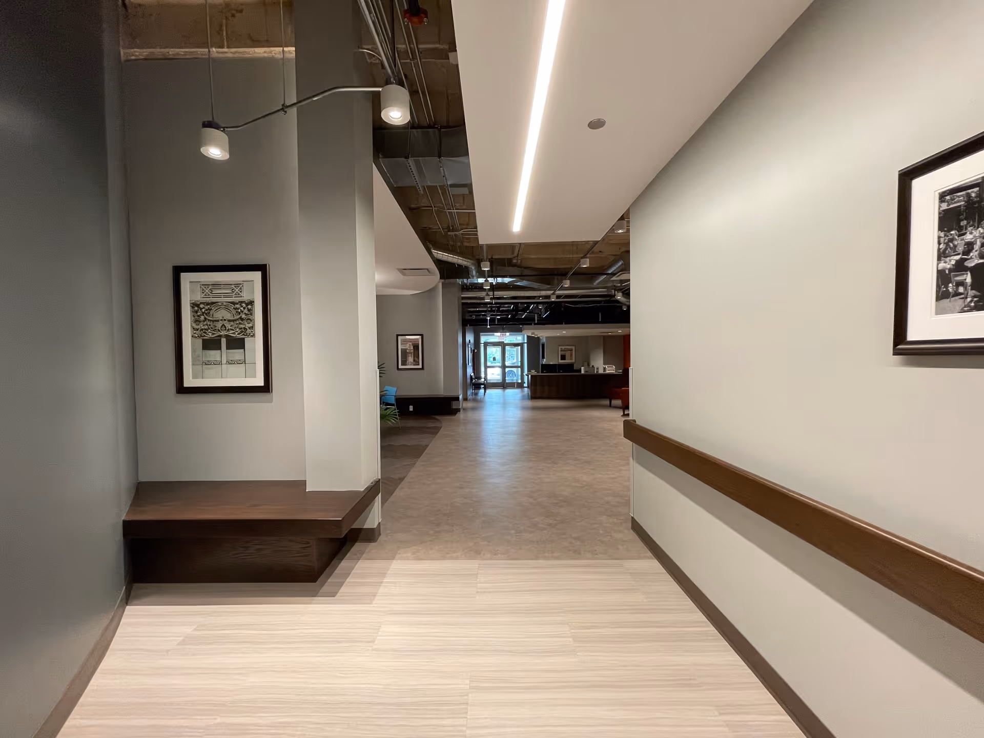 Wide interior hallway with handrails, framed artwork, recessed lighting, and a reception area visible in the distance.