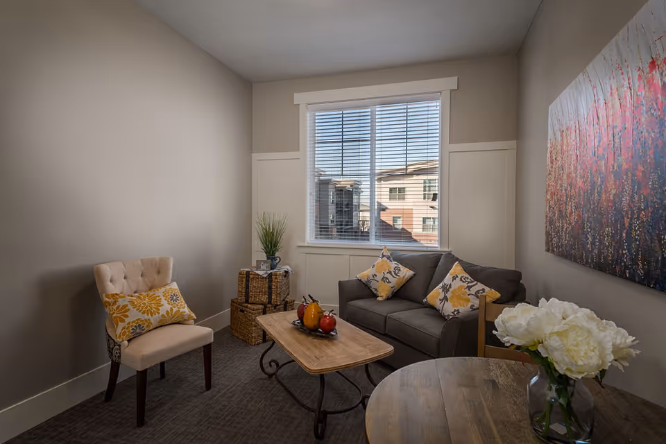 A cozy living room with a gray sofa adorned with yellow and gray floral pillows, a beige upholstered chair with a matching floral pillow, a wooden coffee table with decorative fruit, a round wooden table with a vase of white flowers, and a large colorful abstract painting on the wall. A window with blinds shows a view of neighboring buildings.