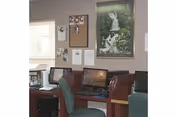 Interior view of a small office or computer area with two computer workstations, green office chairs, a bulletin board with pinned papers and photos, and a framed picture of a statue on the wall.
