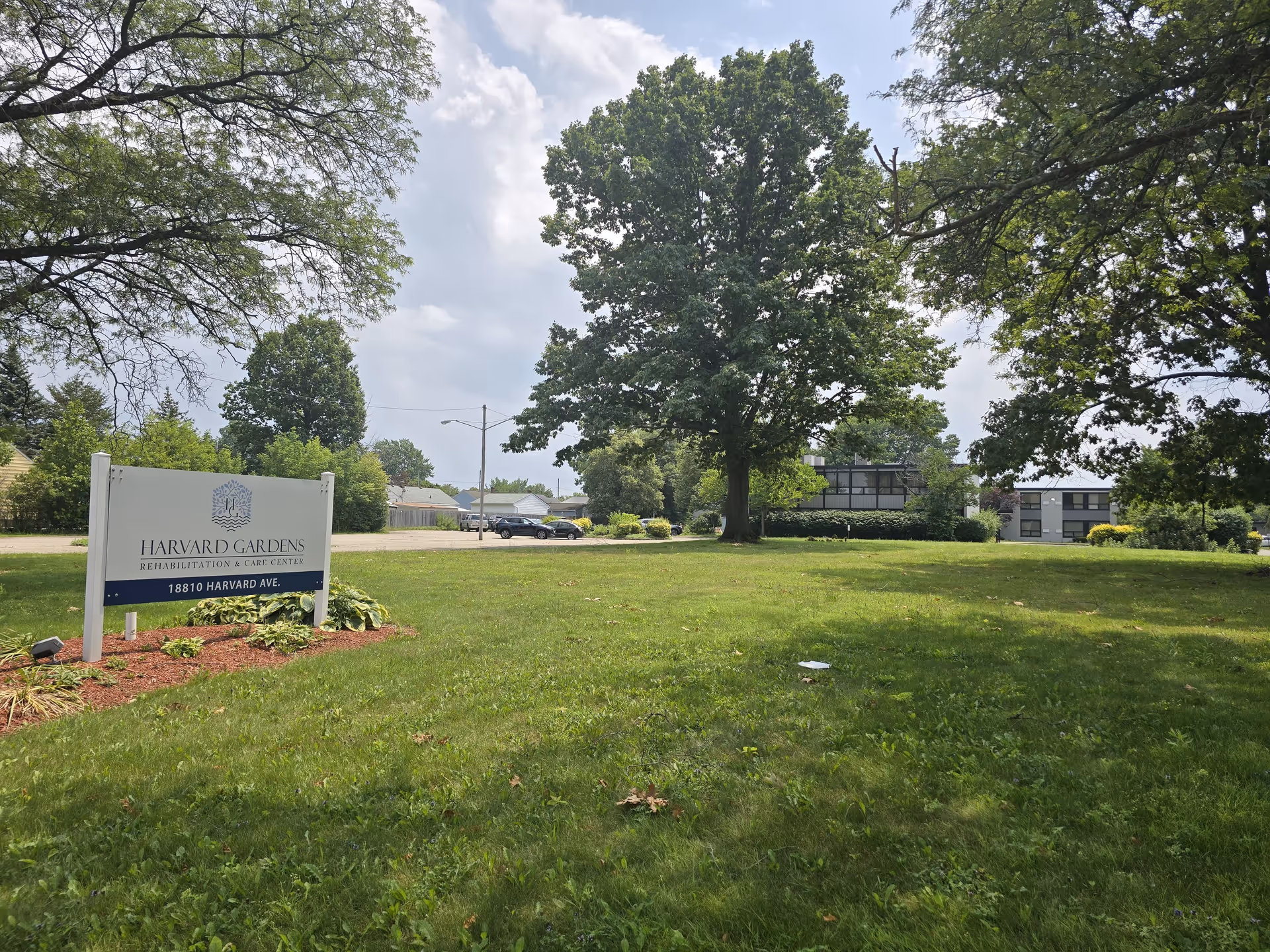 A grassy outdoor area with large trees and a sign that reads 'Harvard Gardens Rehabilitation & Care Center, 18810 Harvard Ave.' In the background, there is a building partially visible behind the trees and some parked cars.