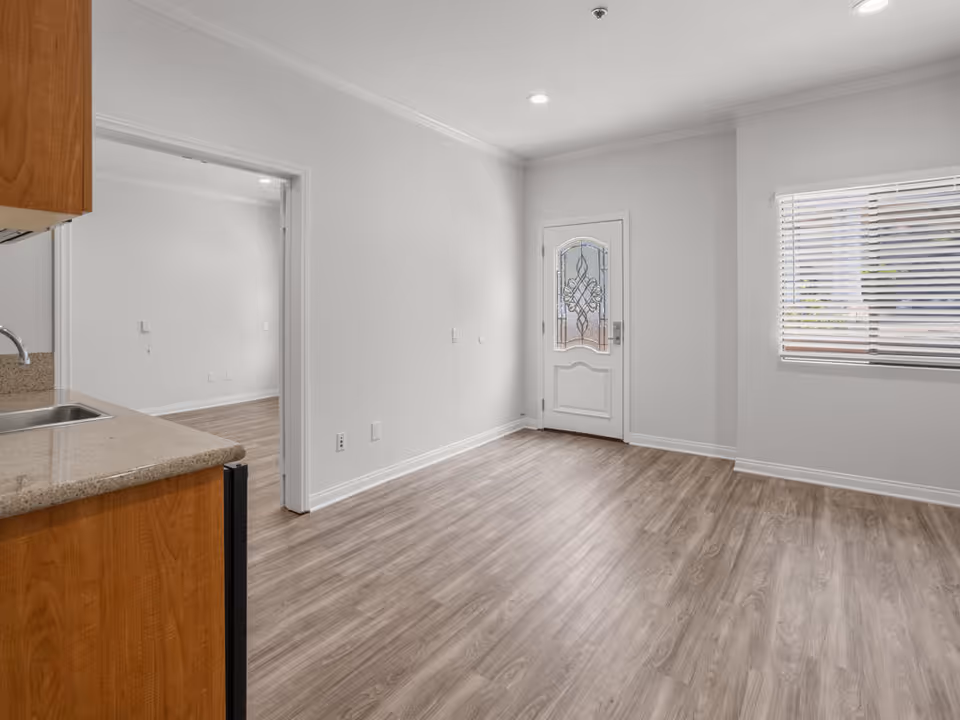 Empty interior room with light wood flooring, white walls, a decorative glass front door, a window with blinds, and a partial view of a kitchen countertop and sink on the left side.