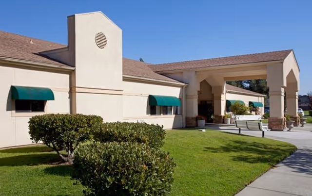 Exterior view of Clearwater Healthcare Center building with beige walls, green window awnings, a covered entrance, and neatly trimmed bushes on a sunny day.