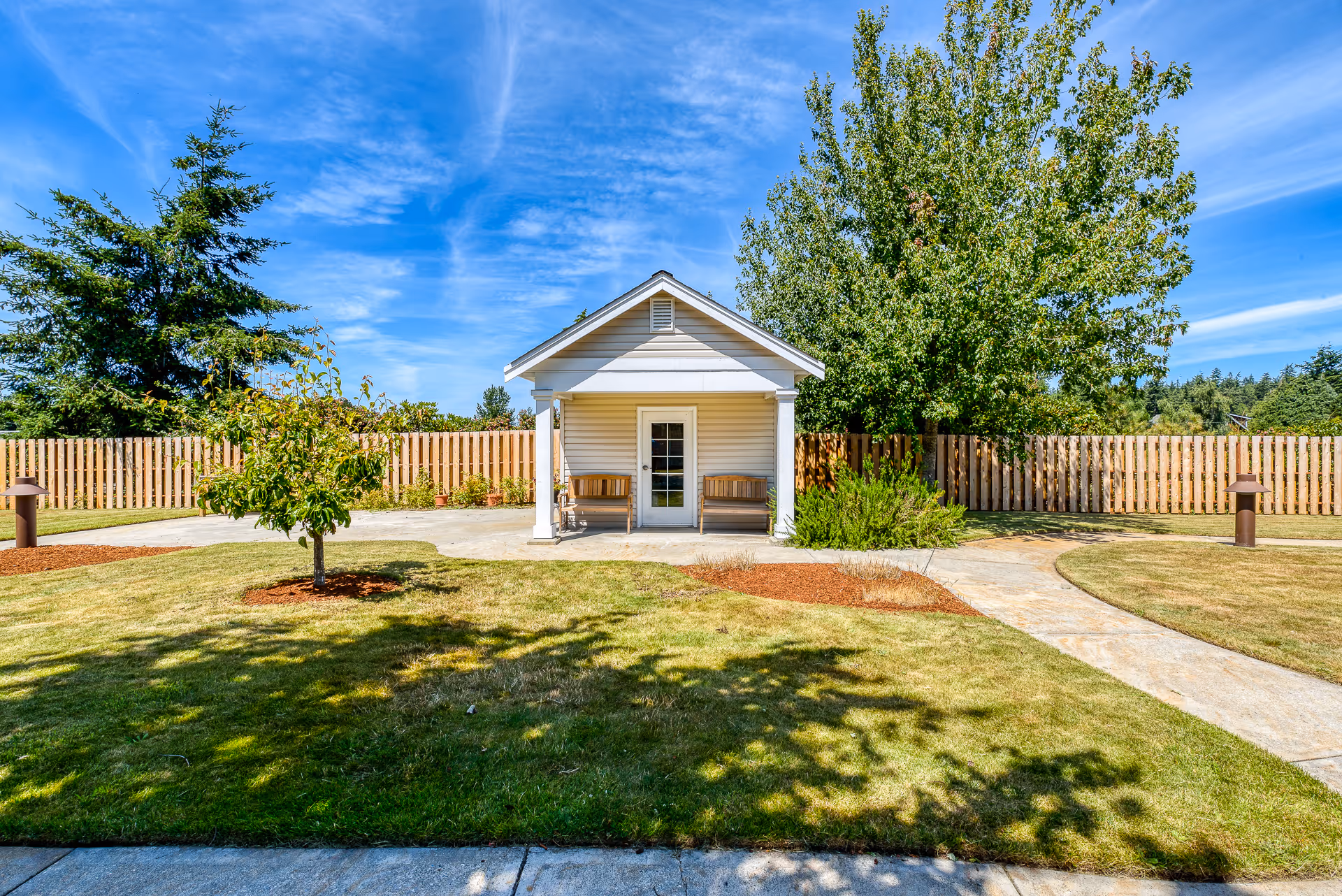 Small white garden pavilion with two benches in a fenced grassy courtyard under a blue sky.