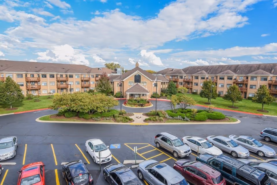 Exterior view of Bridgeway of Bensenville Supportive Living facility showing a large three-story building with balconies, surrounded by landscaped greenery and a parking lot filled with cars under a partly cloudy sky.
