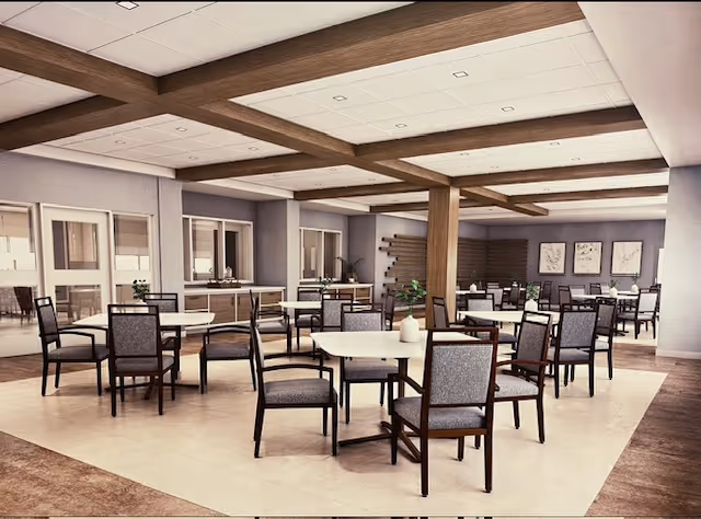 A spacious dining room with multiple square tables and cushioned chairs arranged neatly. The room features wooden ceiling beams, large windows, and neutral-colored walls with framed artwork. There are plants and decorative elements placed around the room, creating a welcoming atmosphere.