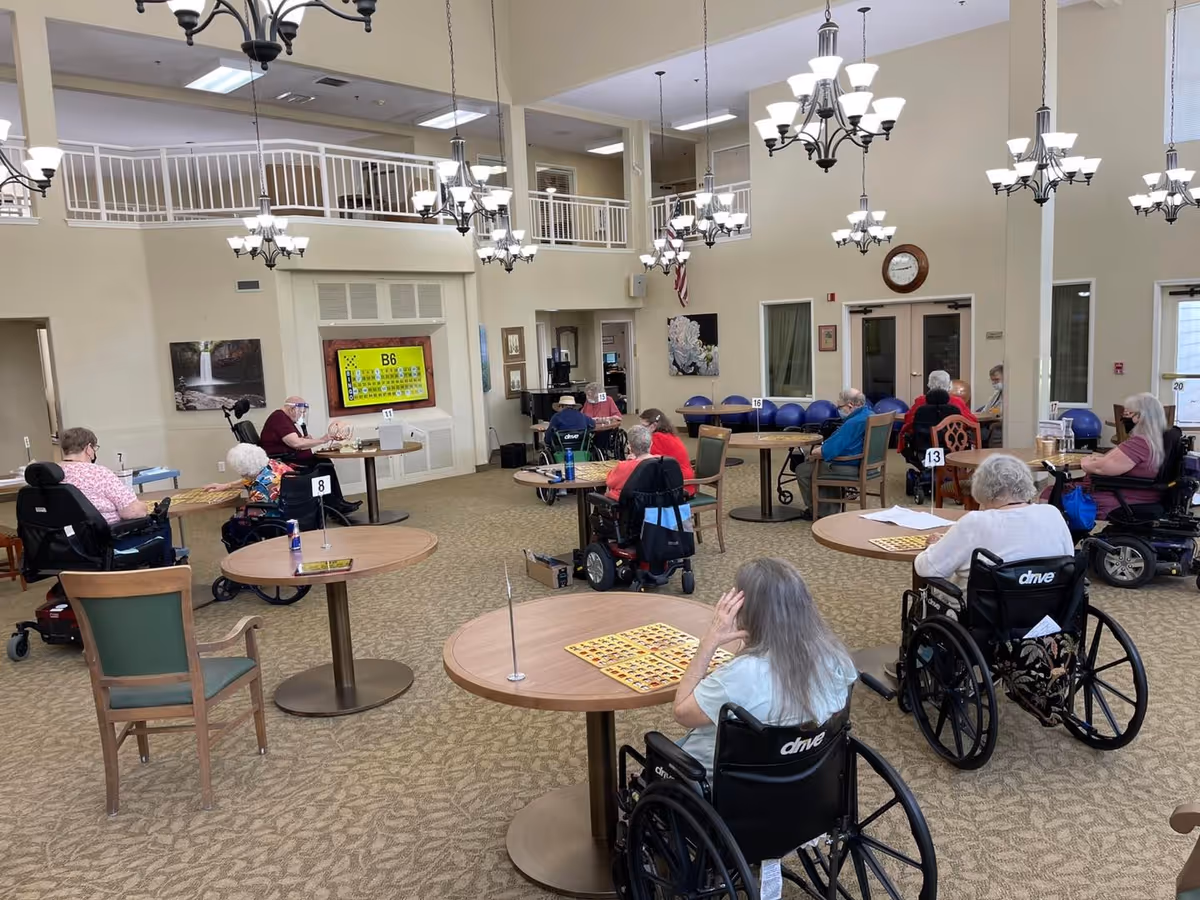 Seniors in wheelchairs seated at round tables in a spacious, well-lit common/activity room with chandeliers and an upper balcony.