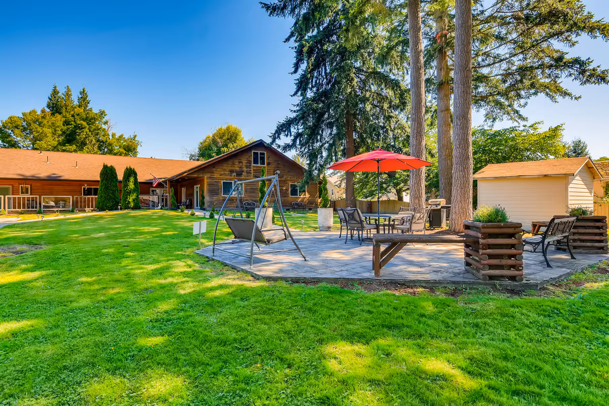Outdoor patio area with a swing chair, table with chairs under a red umbrella, benches, and large trees, set on a paved surface surrounded by green grass. In the background, there is a wooden building with a porch and an American flag.