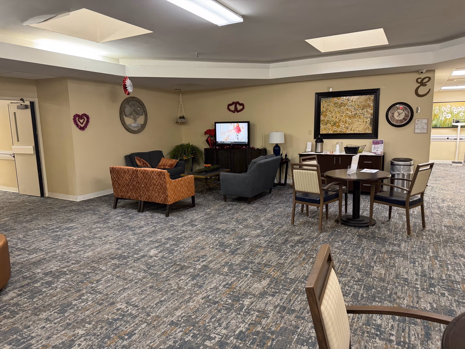 A senior living facility common area with a seating arrangement including a blue loveseat, an orange armchair, and a gray armchair around a coffee table. A television is on a cabinet against the wall, and there is a small round table with four chairs nearby. The walls are decorated with a large framed artwork, a clock, and some heart-shaped decorations. The carpet has a textured pattern, and the ceiling has recessed lighting and skylights.