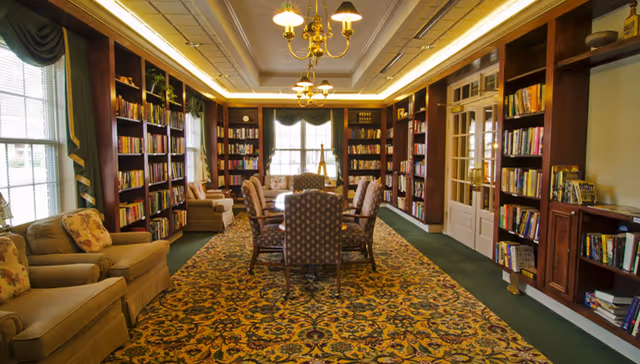 Well-lit library-style common room with bookshelves lining the walls, a central table with chairs, and upholstered seating by the windows.
