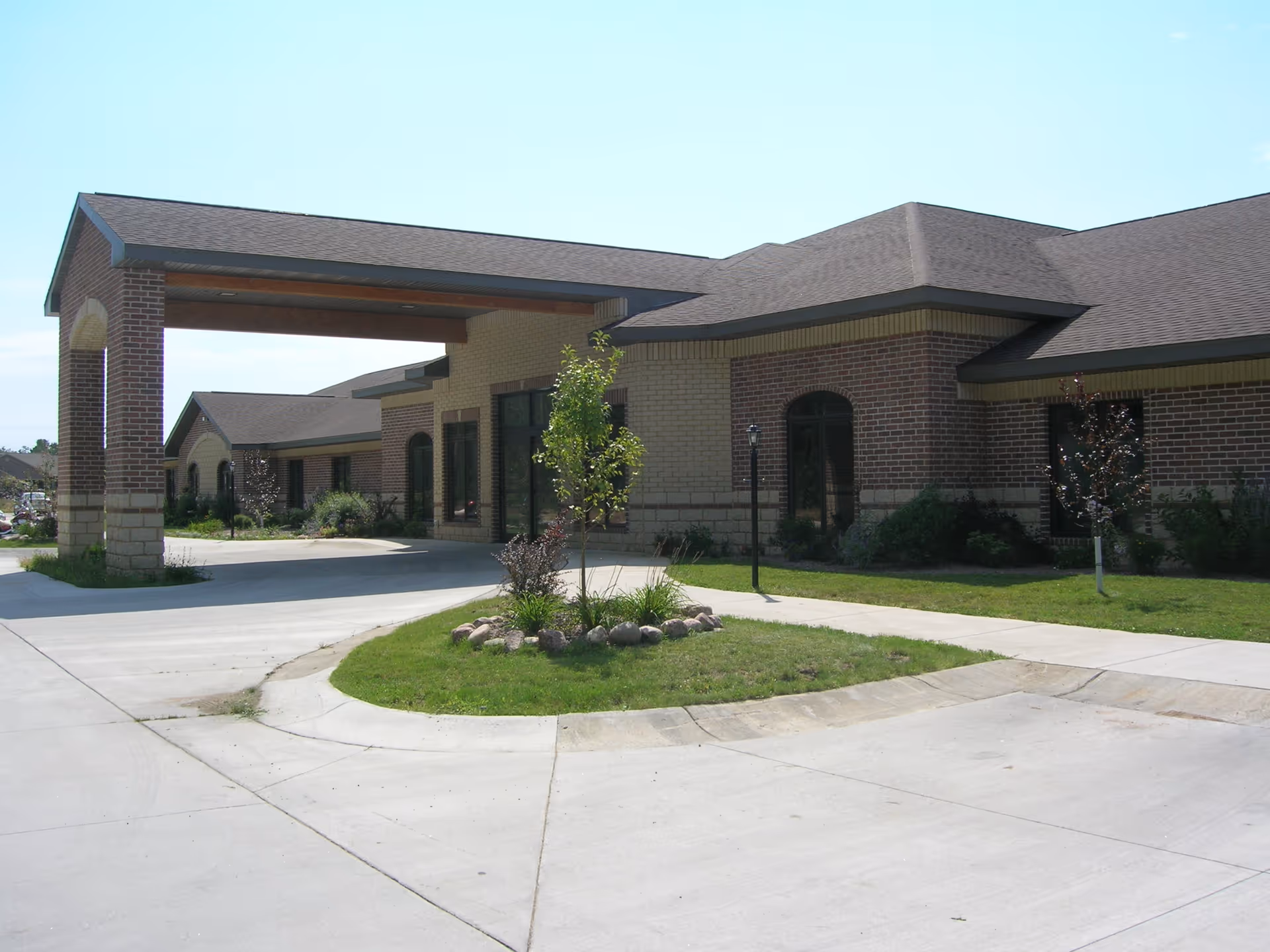 Exterior view of Arborview Court facility showing a single-story brick building with a covered entrance driveway, small landscaped area with grass, plants, and young trees, and a clear blue sky.