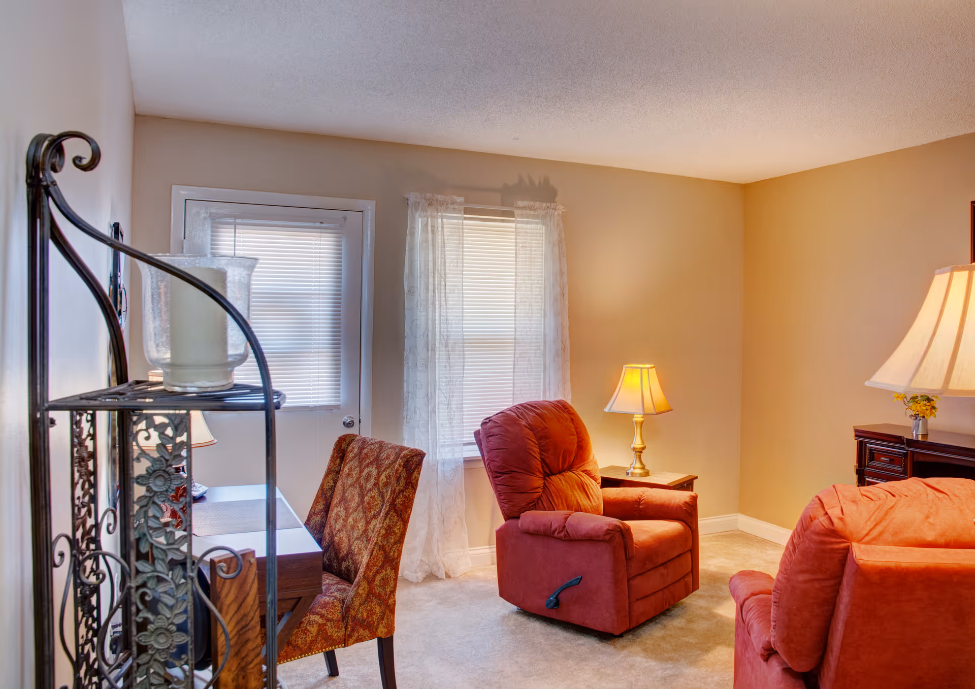 Cozy living room featuring two red recliners, a patterned chair by a small desk, table lamps, and a window with sheer curtains.
