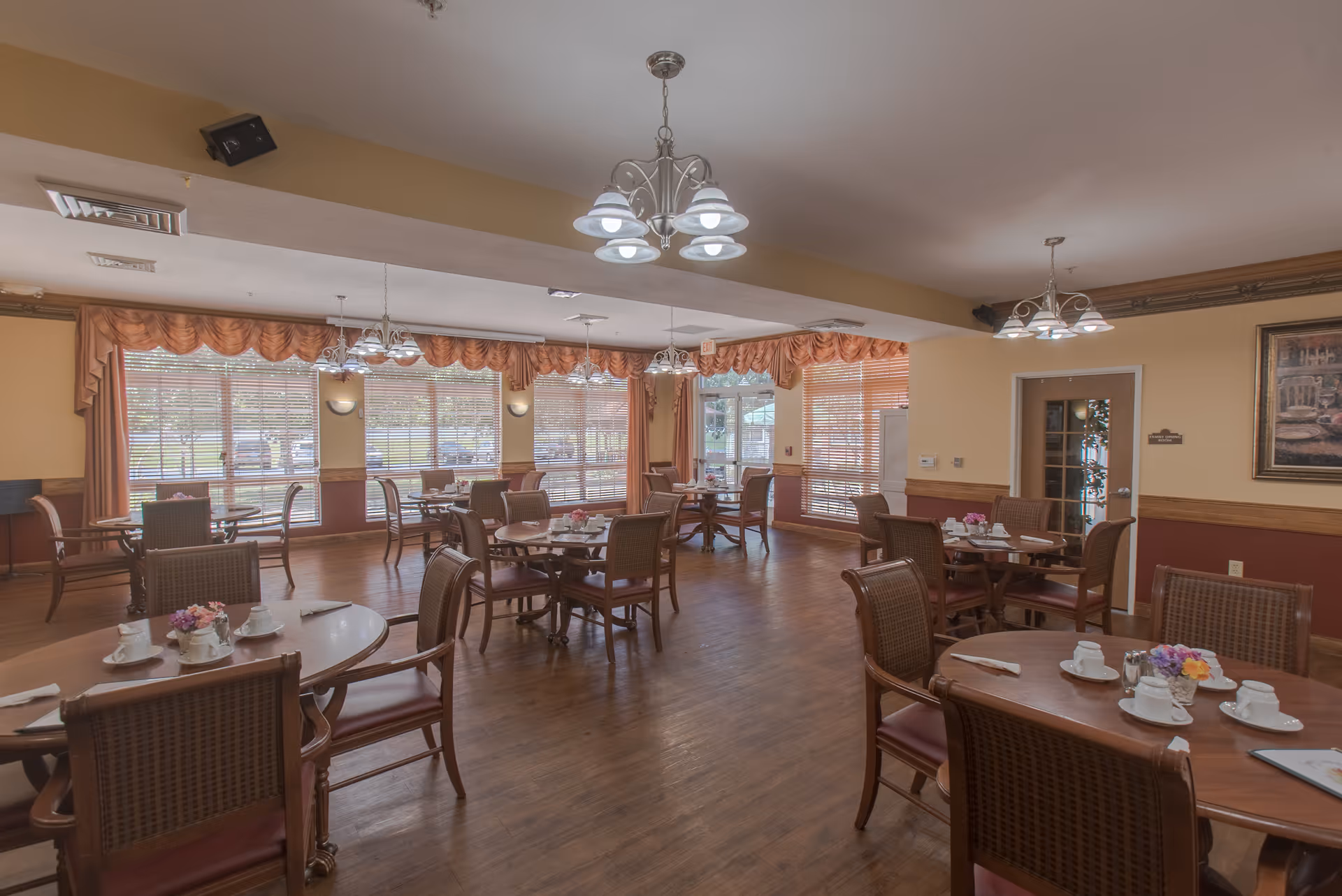 A spacious dining room with multiple wooden tables and chairs arranged neatly. Each table is set with white cups, saucers, napkins, and small flower arrangements. Large windows with peach-colored valances and blinds allow natural light to fill the room. The walls are painted in warm tones with wood trim, and there are several ceiling light fixtures providing additional lighting.