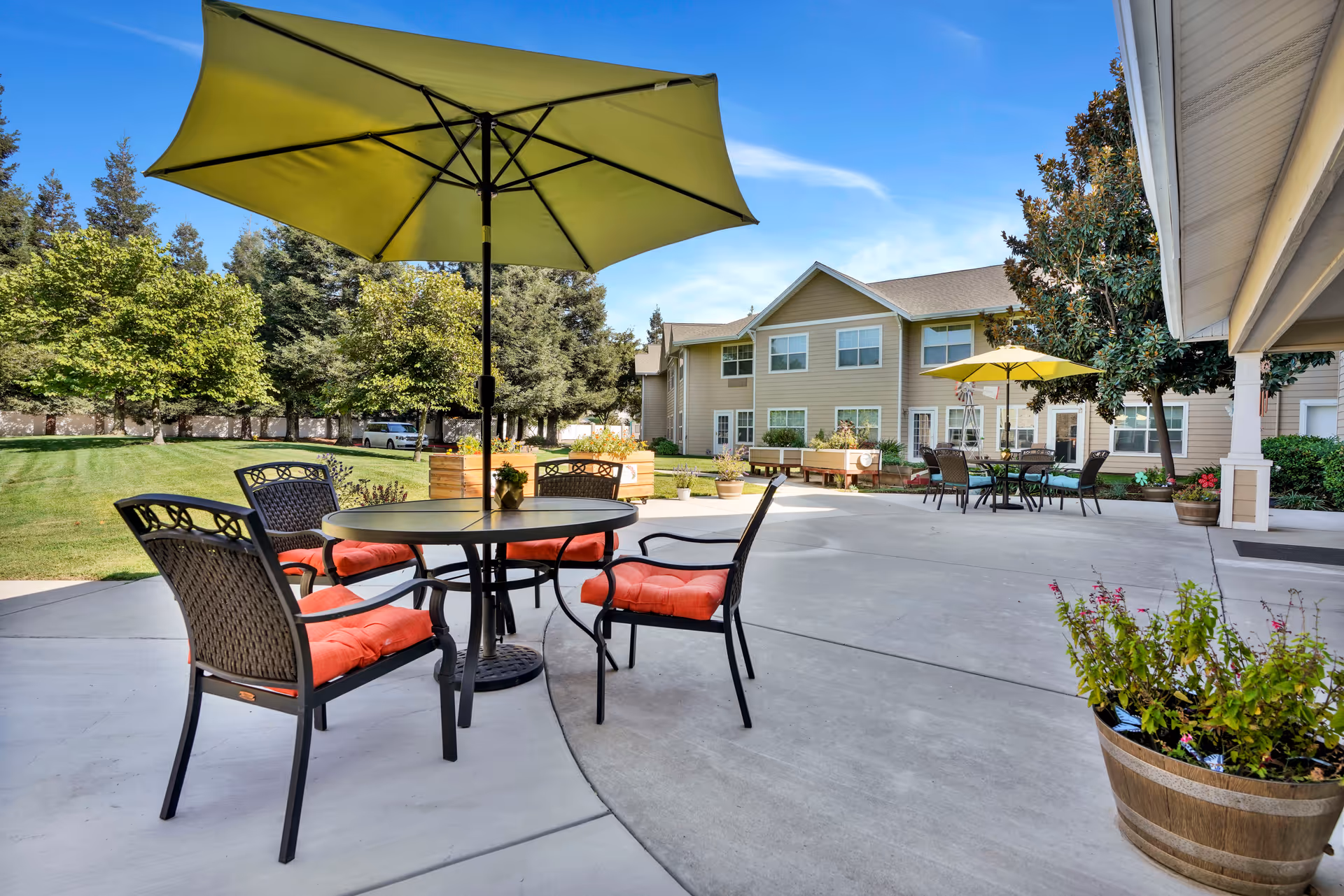 Outdoor patio with tables, chairs and green umbrellas in front of a two-story residential building and landscaped lawn.