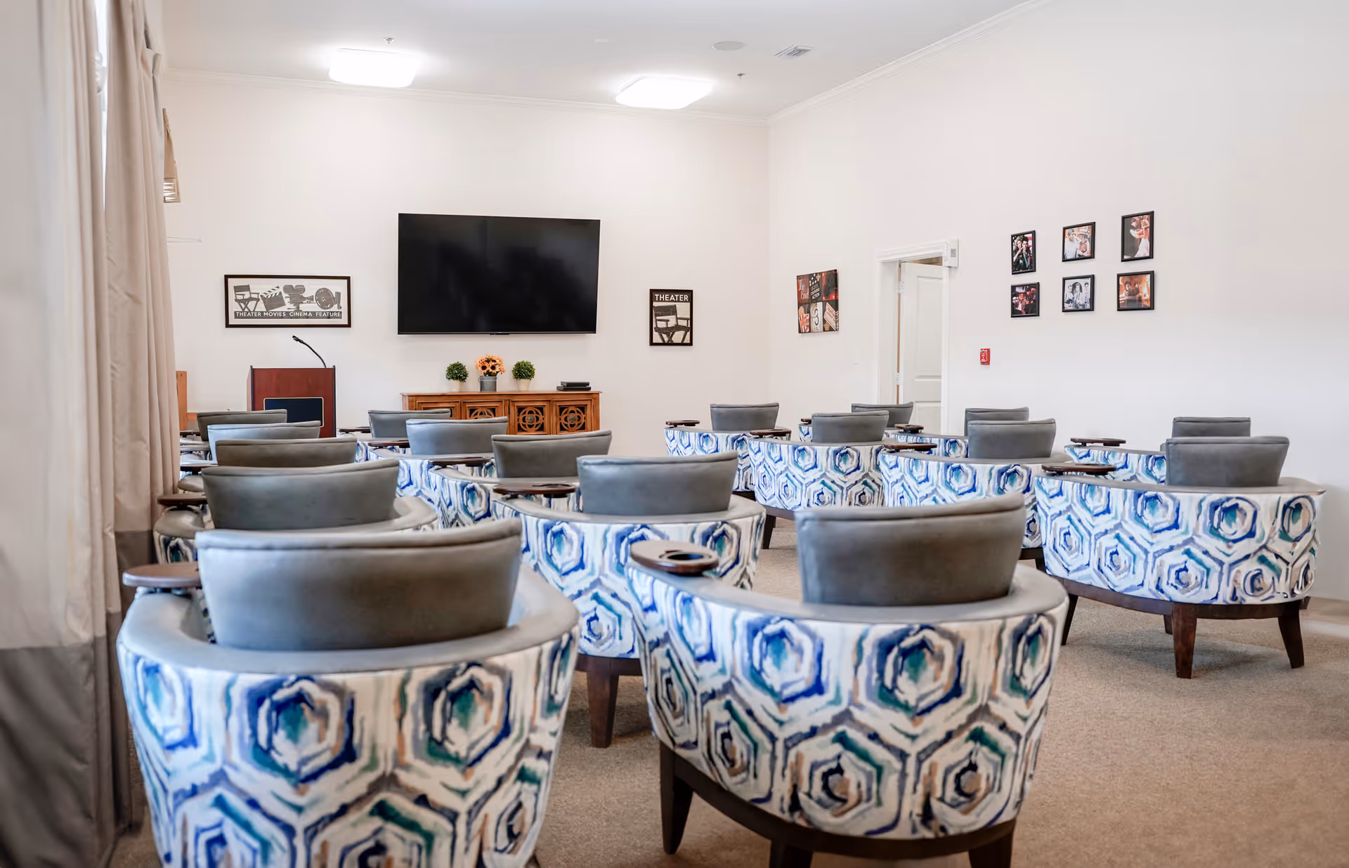 A bright room with multiple patterned armchairs arranged in rows facing a large flat-screen TV mounted on the wall. The room has light-colored walls decorated with framed pictures and a wooden cabinet beneath the TV with small plants and decorative items. There is a podium with a microphone on the left side of the room.