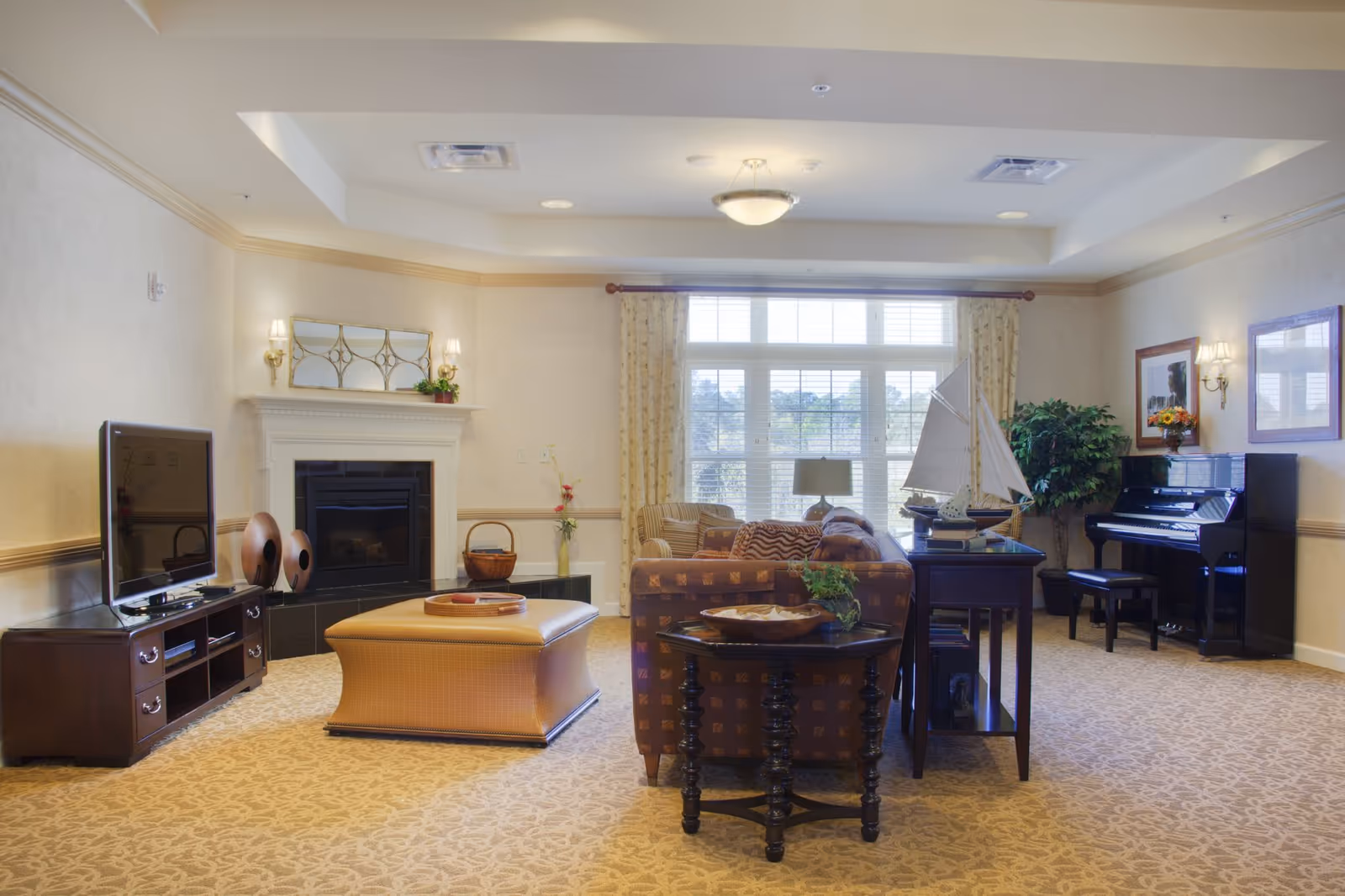 A cozy living room with a patterned carpet, a brown patterned sofa, a large ottoman, a flat-screen TV on a wooden stand, a fireplace with decorative items on the mantel, a large window with curtains, a piano with a bench, and a model sailboat on a side table.