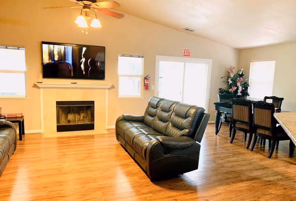 A bright communal living room with leather sofas, a wall-mounted TV above a fireplace, and a dining table in the background.