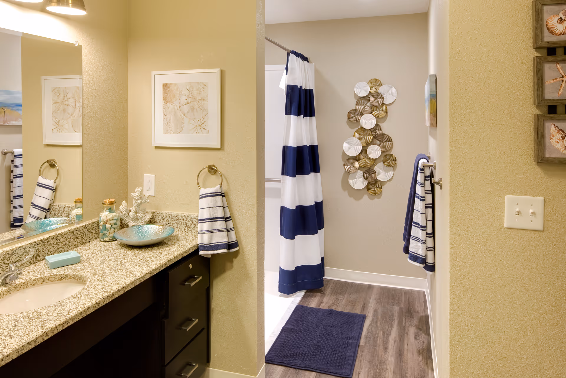 A bathroom with a granite countertop, sink, and dark wood cabinets. There are decorative items on the counter including a bowl and a jar with shells. A large mirror is above the sink. The bathroom has beige walls and a striped blue and white shower curtain. Blue and white towels hang on towel rings and bars. The floor is wood-style with a blue bath mat. Wall decorations include framed pictures and a circular metal art piece.