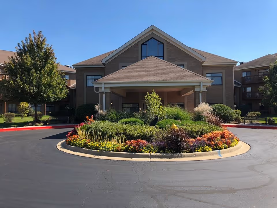 Front exterior view of a two-story supportive living facility building with a covered entrance, surrounded by well-maintained landscaping including a circular flower bed with colorful flowers and green shrubs, and trees on either side under a clear blue sky.