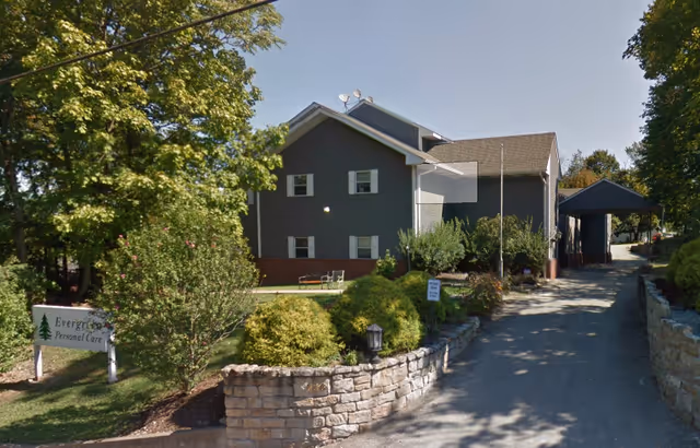 Exterior view of Evergreen Assisted Living facility showing a two-story gray building with white trim, surrounded by trees and bushes. A stone retaining wall lines the driveway leading to a covered entrance on the right. A sign reading 'Evergreen Personal Care' is visible on the left side near the greenery.