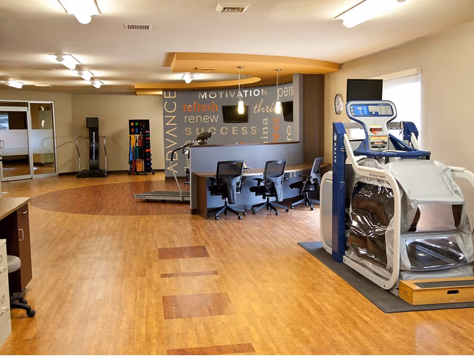A spacious rehabilitation or therapy room with exercise equipment including a treadmill and a large medical machine. There are three office chairs behind a counter, a wall with motivational words, and bright overhead lighting. The floor is wood-patterned and the room has a clean, organized appearance.