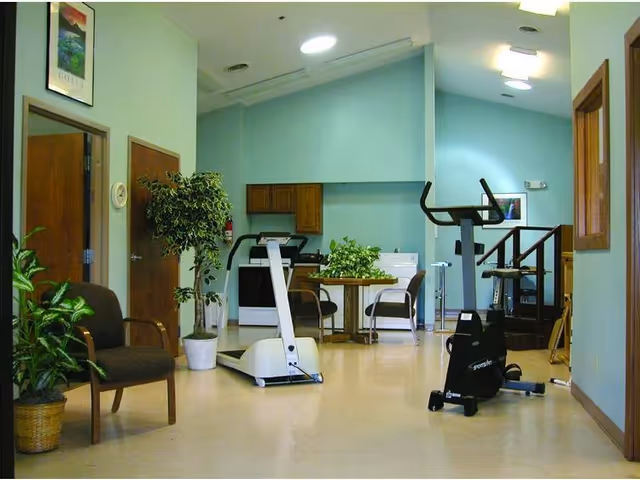Interior room with exercise equipment including a stationary bike and a treadmill, a small table with chairs, potted plants, and a kitchenette area with cabinets and a stove in the background.