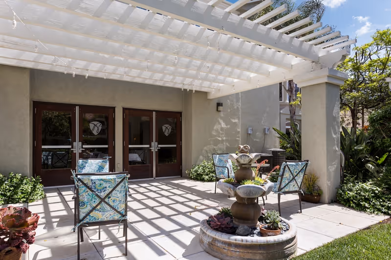 Shaded outdoor patio with a white pergola, a tiered fountain, and patterned chairs arranged in front of double wooden doors.