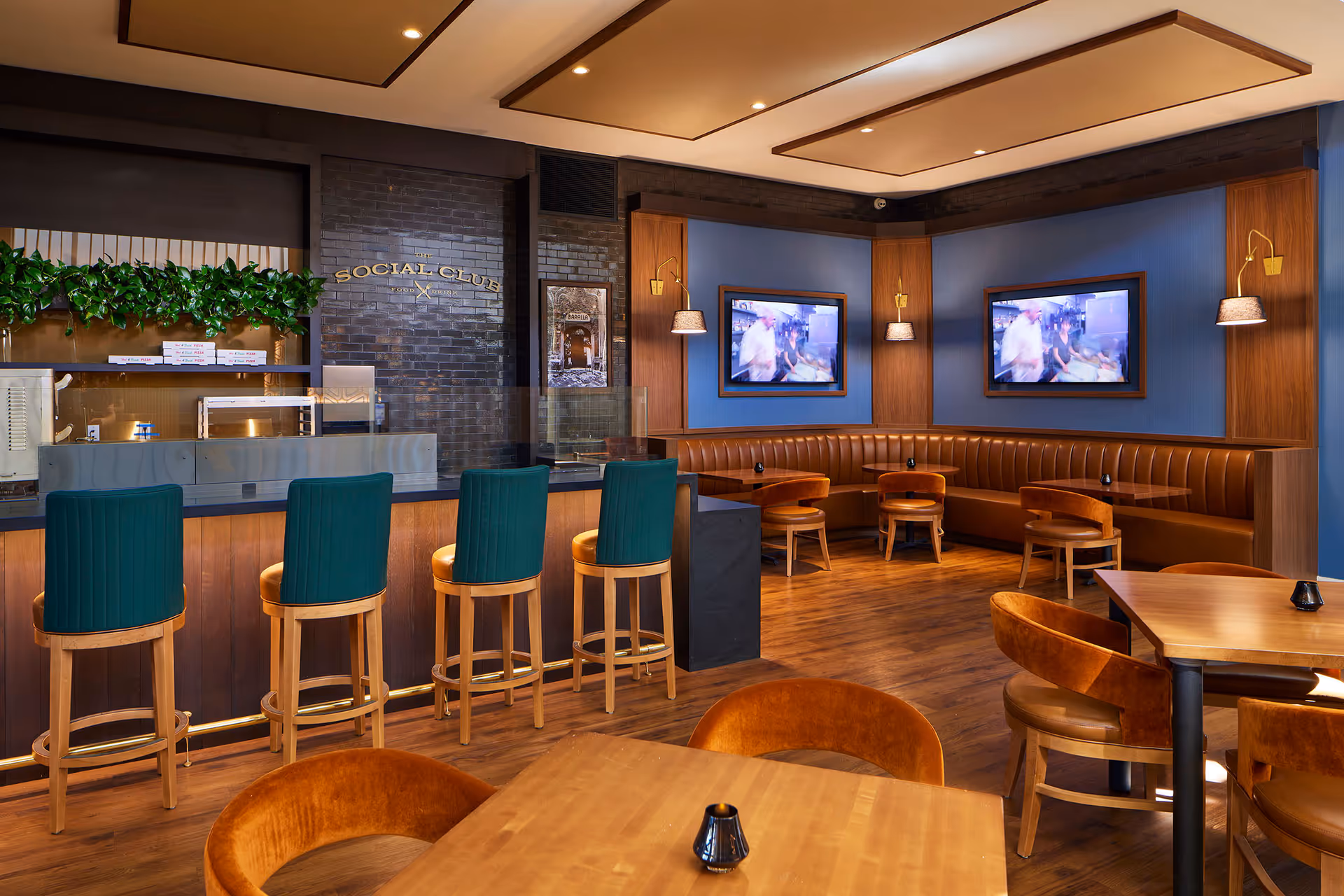 Modern social club dining area with a bar, green-barstools, leather booths, wooden tables and wall-mounted TVs.