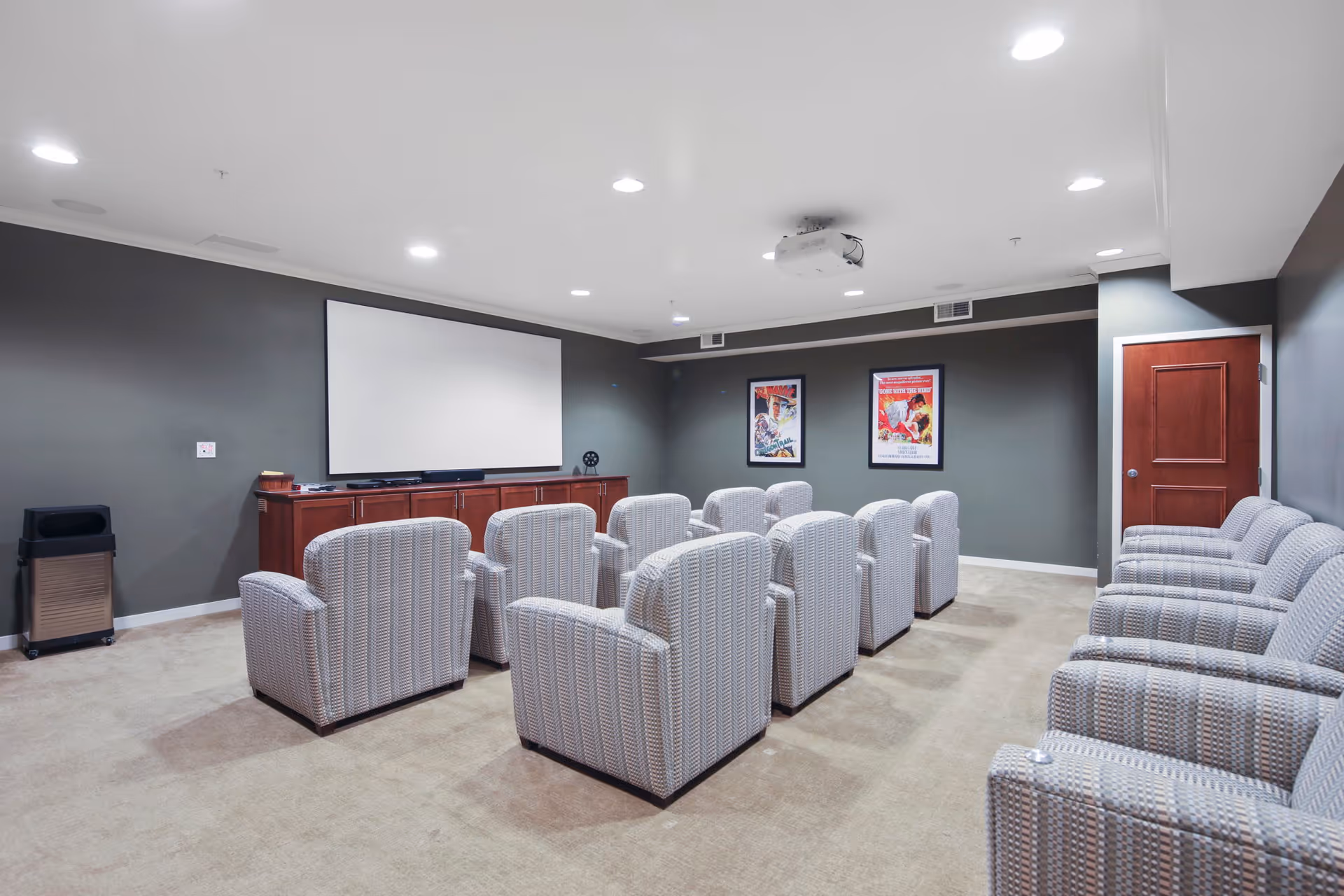 A small movie theater room with two rows of comfortable upholstered armchairs facing a large blank projection screen. The walls are painted gray and decorated with two framed movie posters. There is a wooden cabinet beneath the screen and a projector mounted on the ceiling. A closed wooden door is visible on the right side of the room.