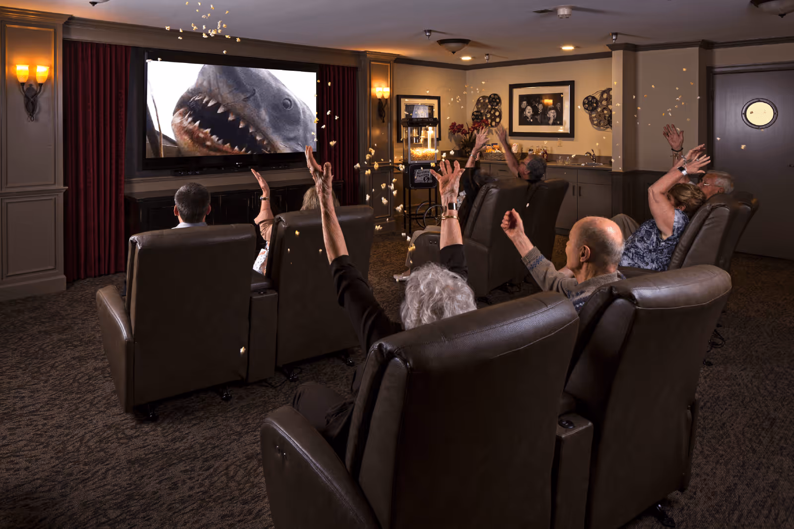 A group of elderly people sitting in comfortable recliner chairs in a small theater room watching a movie featuring a shark on the screen. They are throwing popcorn in the air and appear to be enjoying the movie. The room has warm lighting, movie-themed decorations on the walls, and a popcorn machine in the corner.