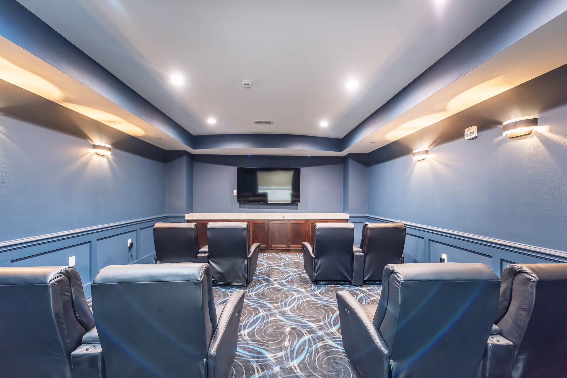 A small theater-style media room with rows of black leather recliners facing a wall-mounted TV in a blue-painted interior.