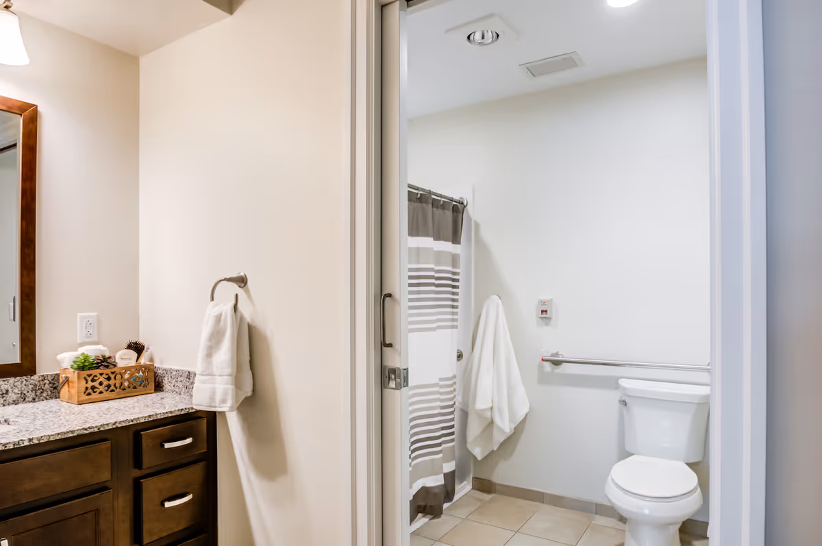 A clean and well-lit bathroom featuring a granite countertop with a wooden cabinet below, a mirror with a wooden frame, a towel hanging on a ring, a shower with a striped curtain, a white towel hanging on a hook, a toilet with a grab bar beside it, and tiled flooring.
