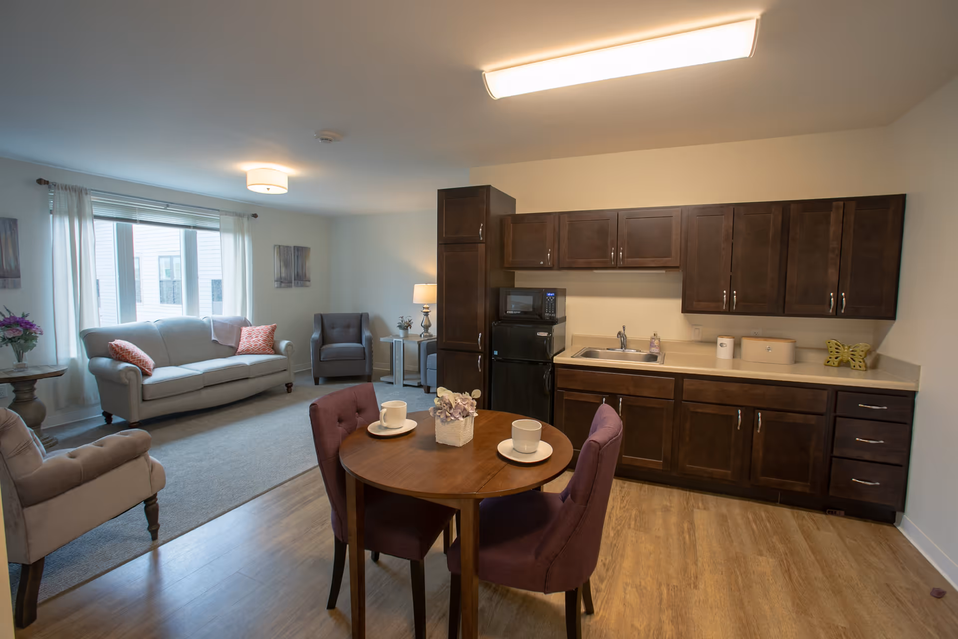 A cozy living area and kitchenette in a senior living facility. The room features a small round wooden dining table with two purple upholstered chairs, set with white cups and saucers. The kitchenette has dark wood cabinets, a small black refrigerator, microwave, and a sink. The living area includes a light gray sofa with pink pillows, a gray armchair, a side table with a lamp, and a window with white curtains letting in natural light.