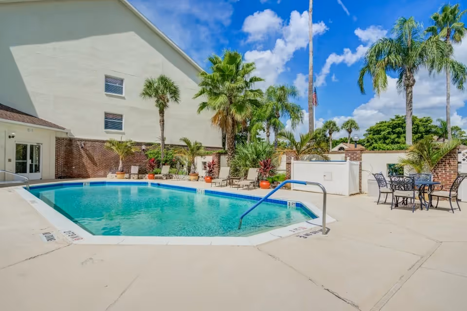 Outdoor swimming pool area at Merrill Gardens at Barkley Place with clear blue water, surrounded by palm trees, lounge chairs, potted plants, and a small table with chairs under a bright blue sky with scattered clouds.