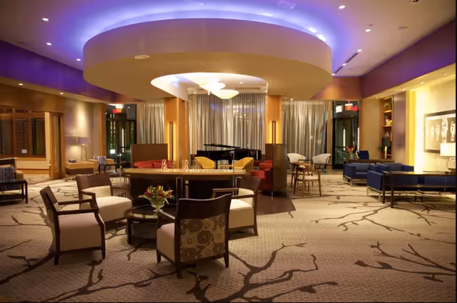 A spacious and elegantly furnished living room area with various seating arrangements including armchairs, sofas, and small tables. The room features a large circular ceiling light with blue accent lighting, a grand piano in the background, and large windows with curtains. The carpet has a tree branch pattern, and the room is warmly lit with lamps and recessed lighting.