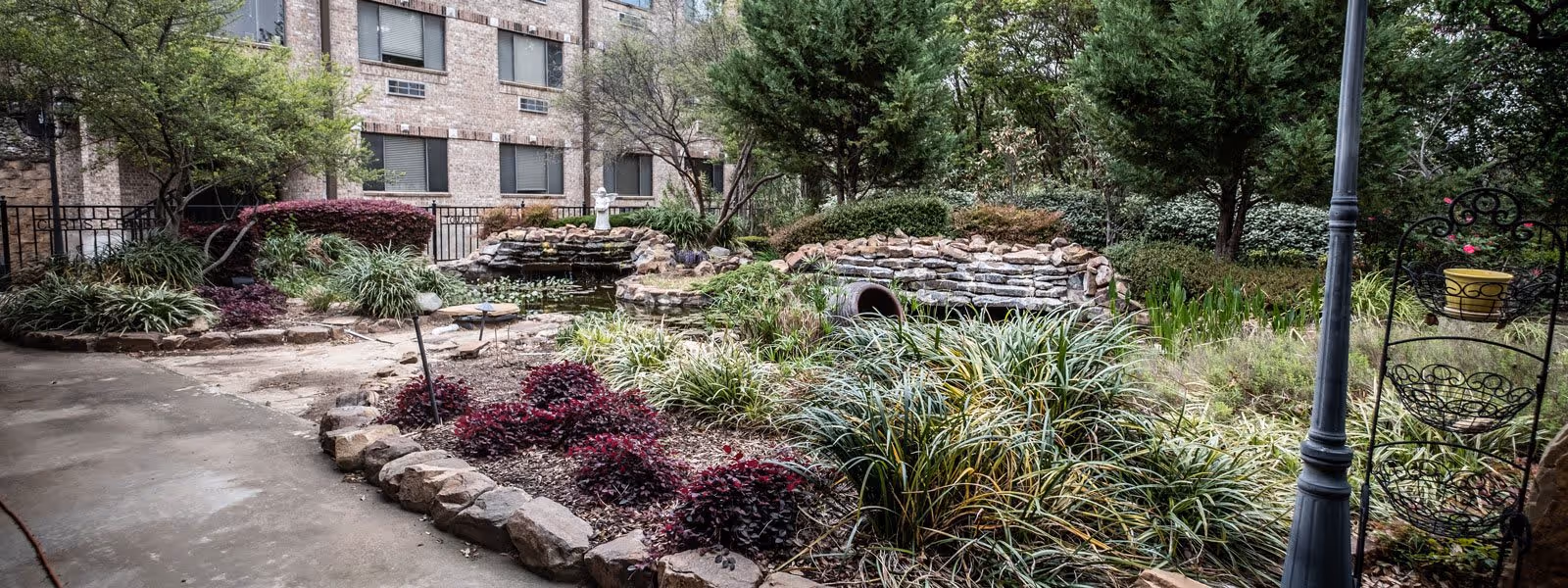 A landscaped outdoor garden area at a senior living facility featuring a stone-bordered pond with a small waterfall, surrounded by various green plants, shrubs, and trees. A brick building with multiple windows is visible in the background. There is a paved walkway on the left side and a decorative metal plant stand with a yellow pot on the right side.