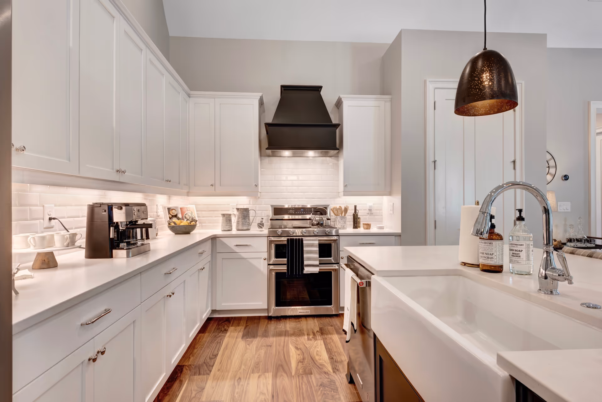 Modern kitchen with white cabinetry, a stainless steel stove with a black range hood, a coffee machine on the counter, a large white farmhouse sink with a chrome faucet, and wooden flooring.