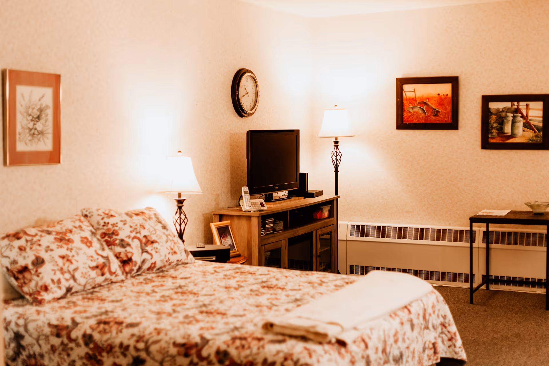A cozy bedroom with a floral patterned bedspread and pillows on a bed. There are two lamps on either side of a wooden TV stand holding a flat-screen television, a cordless phone, and some framed photos. The walls are decorated with framed artwork and a round clock. A small table with a bowl and some papers is placed against the wall.
