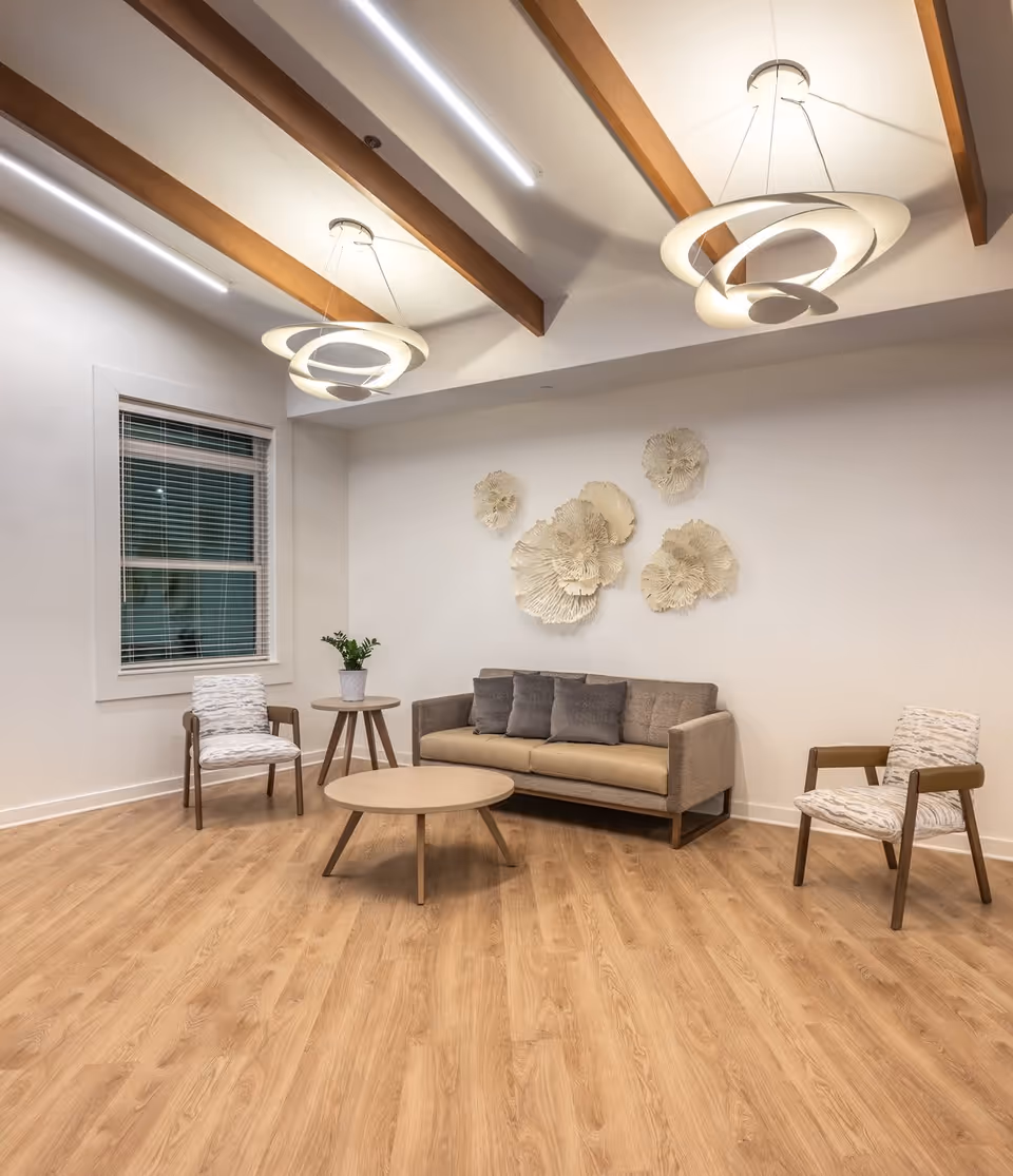 Bright modern lounge with a sofa, two chairs, a round coffee table, decorative wall art, and hanging pendant lights beneath wooden ceiling beams.