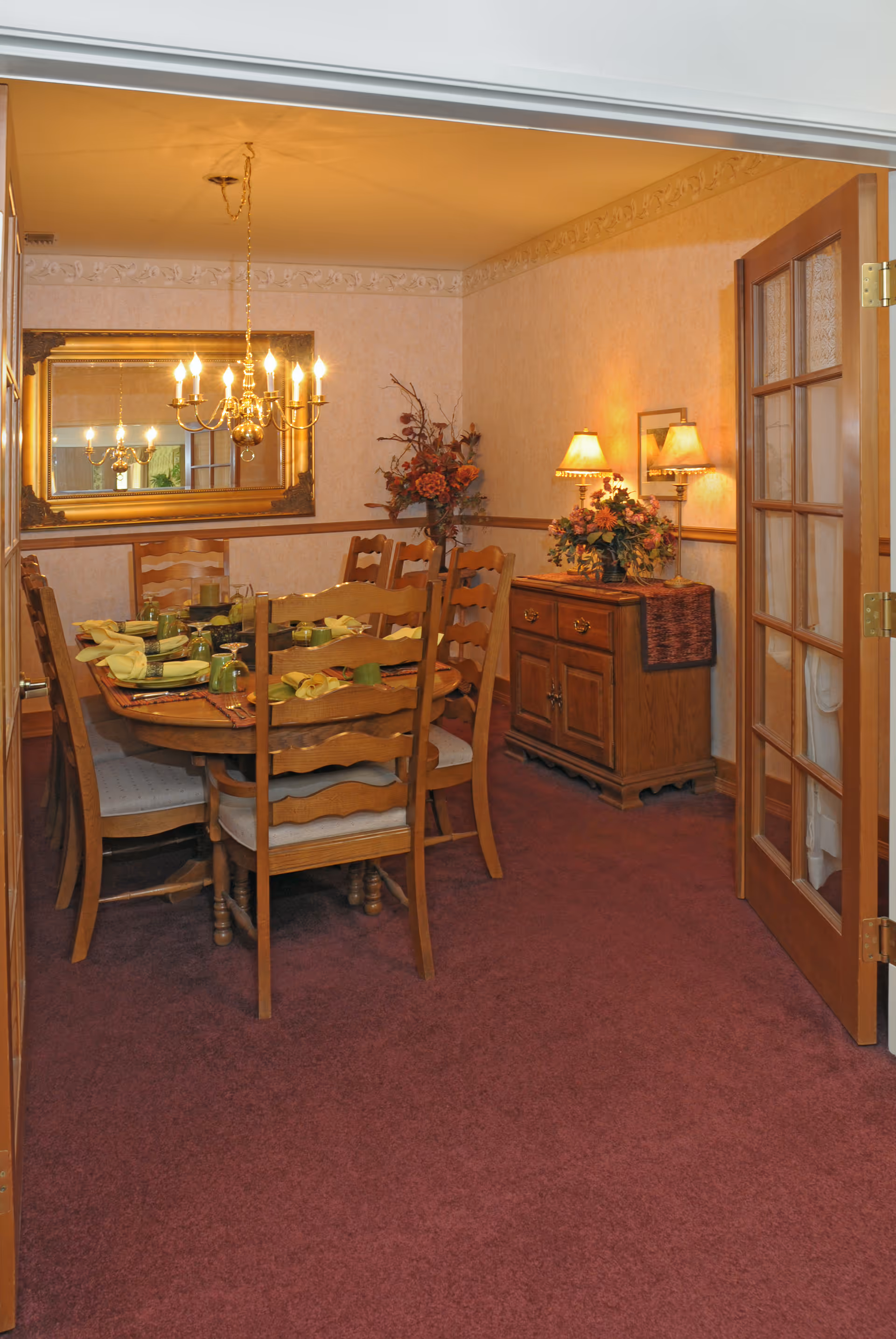 A warmly lit dining room with a wooden dining table set for six people with green glasses and yellow napkins. The room features a large ornate mirror on the wall, a chandelier hanging above the table, a wooden sideboard with floral arrangements and lamps, and an open wooden door with glass panels. The carpet is a deep burgundy color.