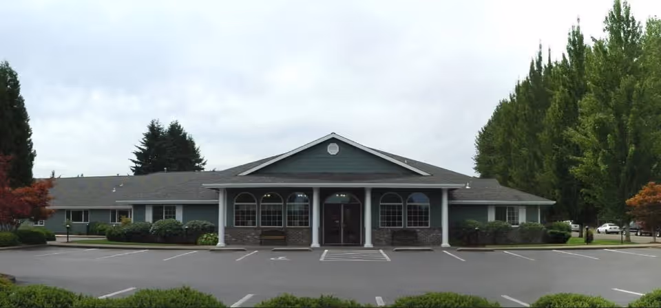 Front exterior view of a single-story building with a covered entrance supported by white columns, surrounded by trees and bushes, with an empty parking lot in front under a cloudy sky.