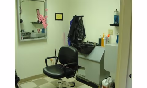 Small room with a black salon chair in front of a hair washing station with a sink. A mirror is mounted on the wall with some pink decorations on it. Various hair care products and cleaning supplies are placed on shelves and counters. The floor has a checkered pattern.
