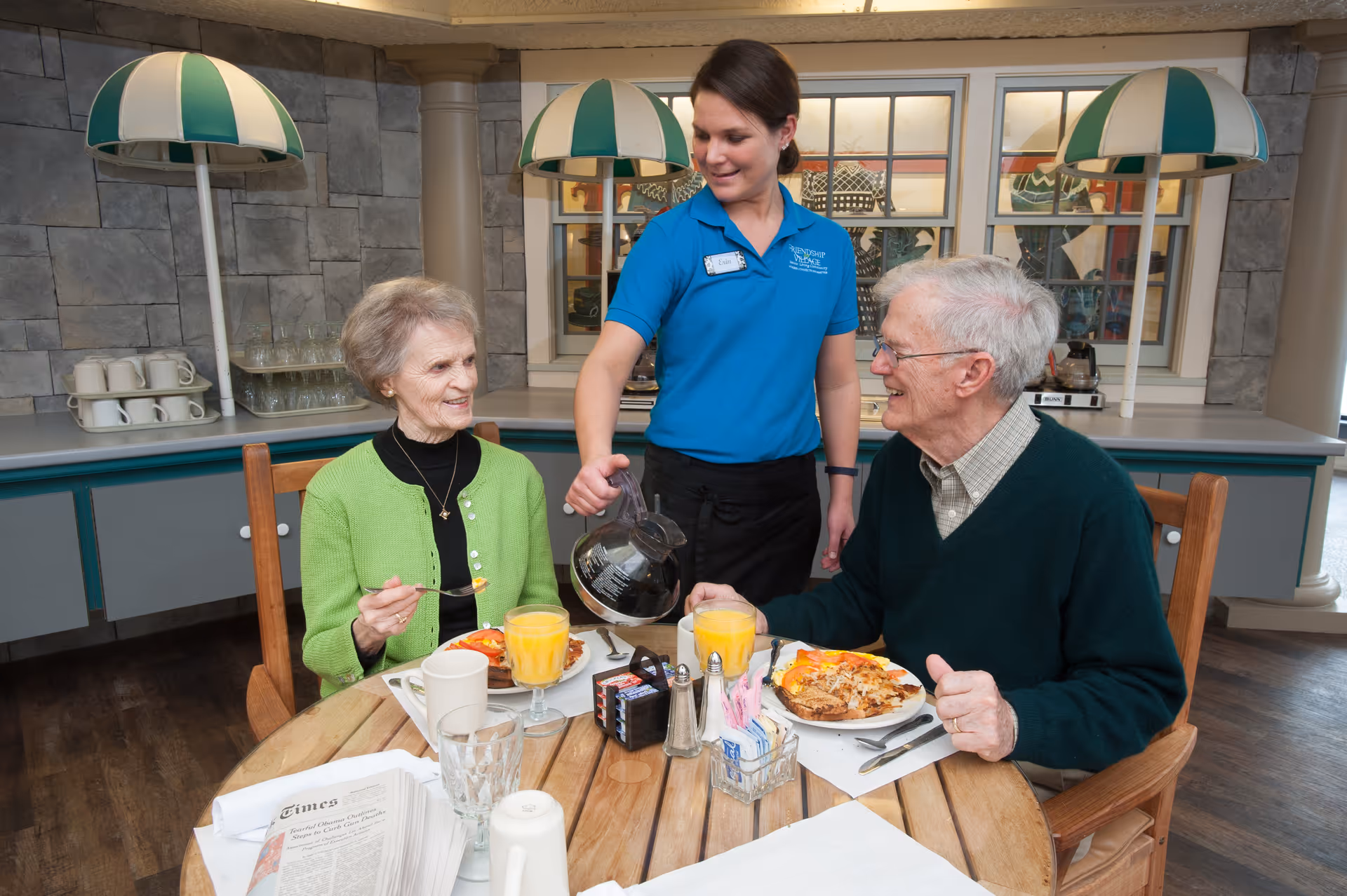 A staff member pours coffee for two elderly residents seated at a round dining table with breakfast plates and orange juice.