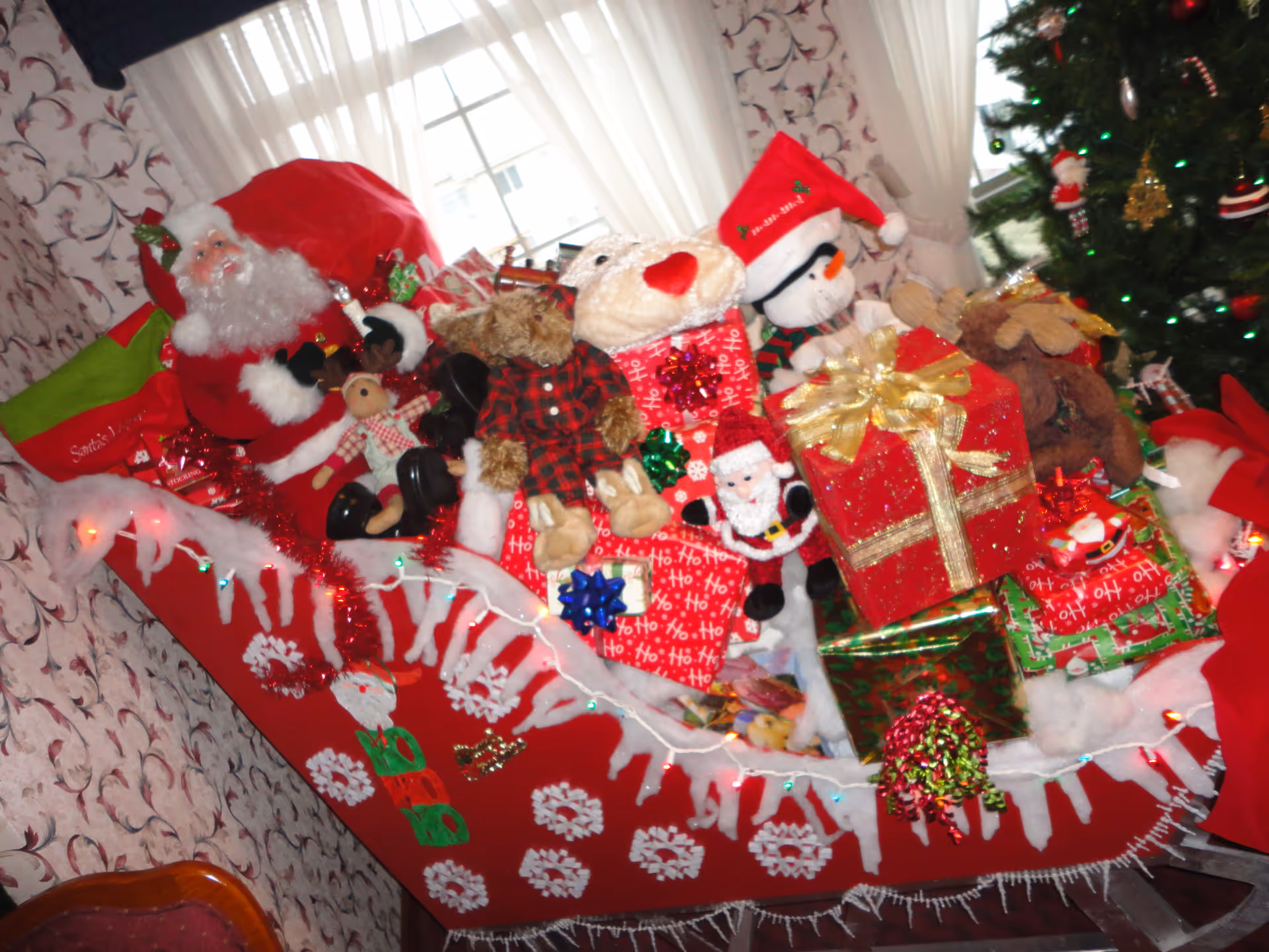 A festive Christmas display featuring a red sleigh filled with wrapped presents, stuffed animals including a teddy bear, a polar bear, a snowman wearing a Santa hat, and a Santa Claus figure. The sleigh is decorated with white faux snow, snowflakes, and colorful lights. In the background, there is a decorated Christmas tree and a window with sheer white curtains.