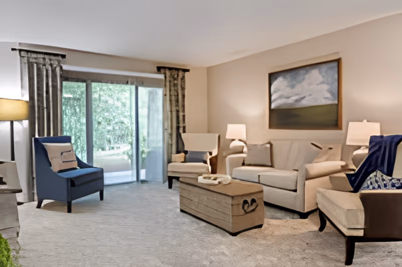 A cozy living room with a beige sofa, two armchairs, a wooden coffee table, and two table lamps. There is a large painting on the wall above the sofa and a sliding glass door with curtains leading to an outdoor area.