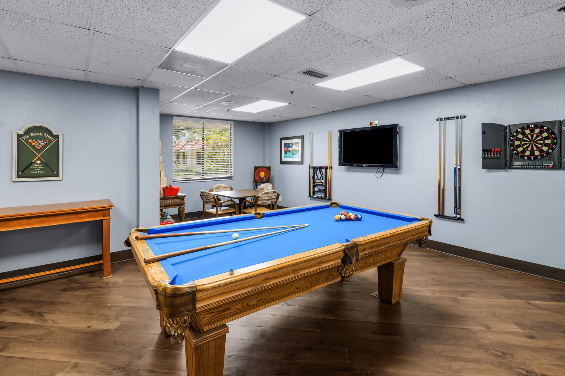 A recreational room with a blue felt pool table in the center, two pool cues resting on the table, a dartboard mounted on the wall, a flat-screen TV, and a small table with four chairs near a window with blinds. The walls are painted light blue and the floor is wooden.