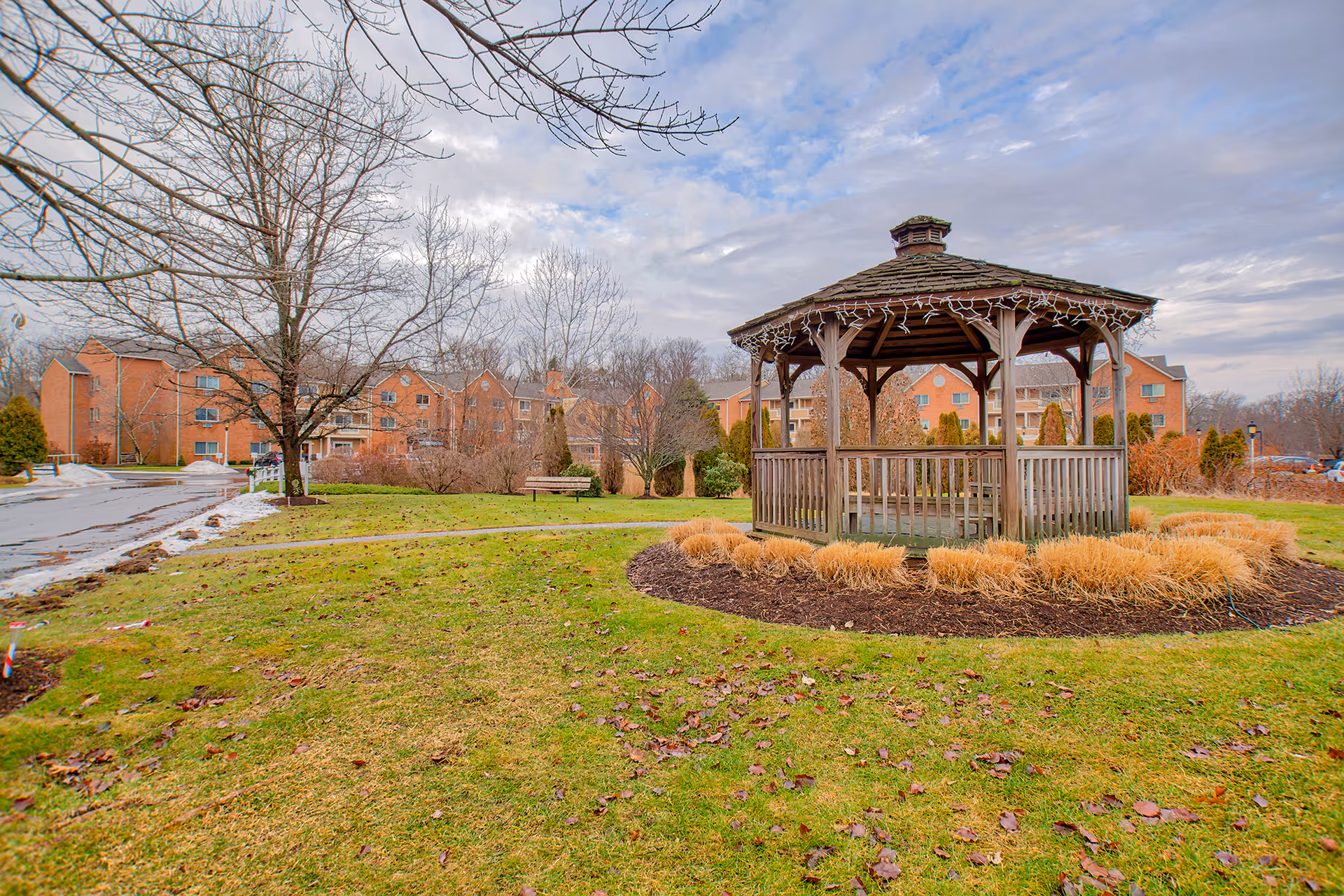 A wooden gazebo with a shingled roof and decorative string lights stands in a grassy area surrounded by landscaping with dried ornamental grasses. In the background, there are leafless trees and a large brick senior living facility building under a partly cloudy sky.