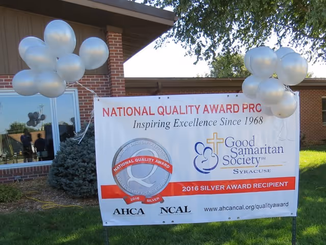 Outdoor scene showing a banner for Good Samaritan Society - Syracuse celebrating their 2016 Silver National Quality Award. The banner is attached to stakes in the grass in front of a brick building with windows. White balloons are tied to the stakes on both sides of the banner.