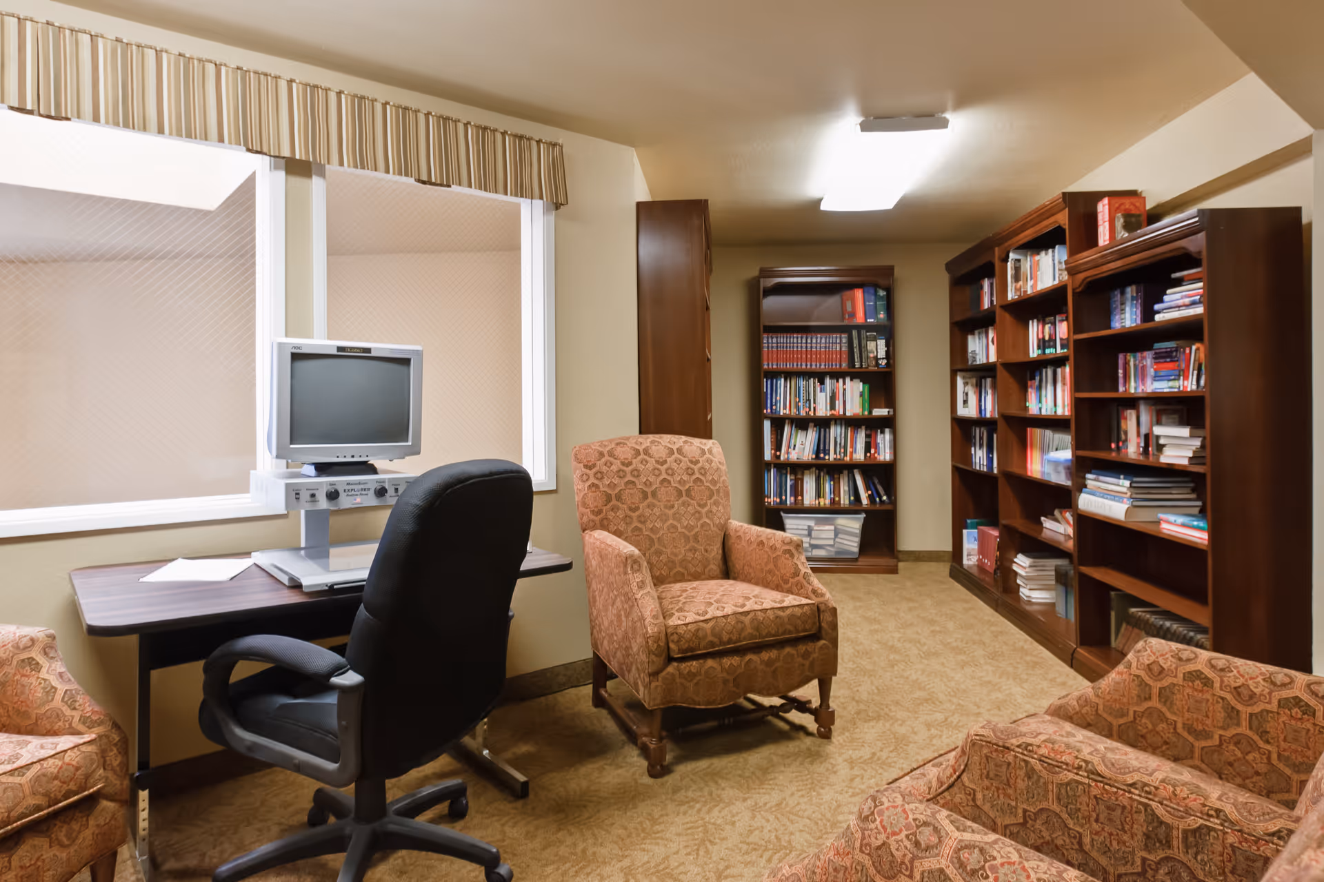 A cozy reading room with upholstered armchairs, bookshelves, and a computer workstation by a window.