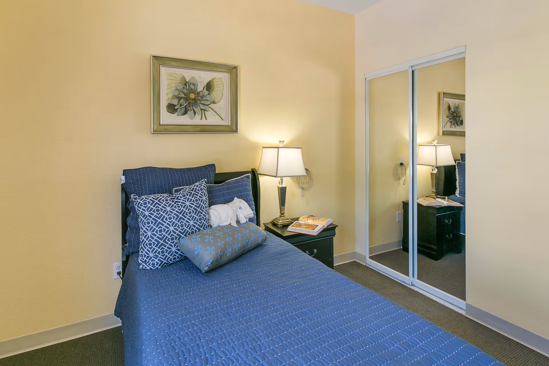 A small bedroom with a single bed covered in a blue quilt and multiple decorative pillows. A bedside table with a lamp, an open book, and reading glasses is next to the bed. There is a framed floral artwork on the wall above the bed and a closet with sliding mirrored doors on the right side.